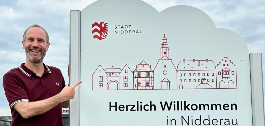Mann stehend neben einem weißen Ortseingangsschild mit Schriftzug "Herzlich Willkommen in Nidderau"