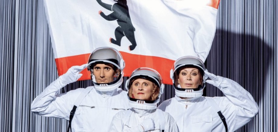 2 Frauen und 1 Mann in weißen Astronautenanzügen vor einer Berliner Flagge