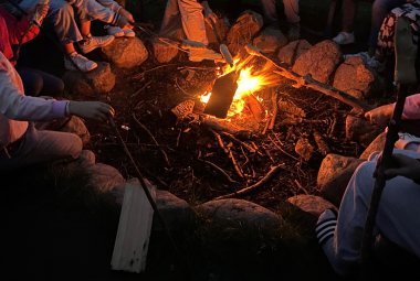 Kinder sitzen um ein Lagerfeuer und halten Stockbrot in das Feuer