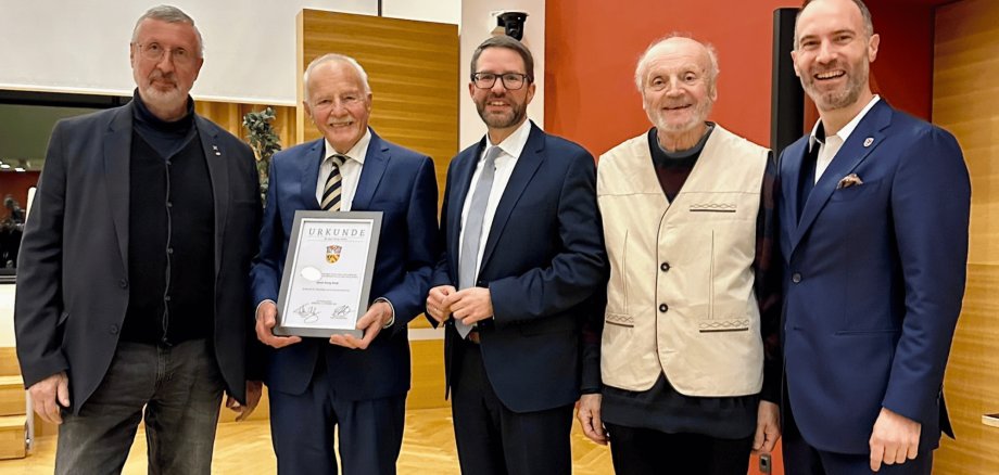 Fünf Männer in Anzügen stehend. Ein Mann hält eine Urkunde vor sich